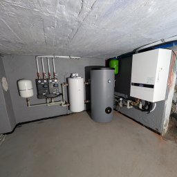 Zestaw urządzeń grzewczych w kotłowni: piec gazowy, zbiorniki, pompy obiegowe i osprzęt. Szare ściany i podłoga, widoczne rury instalacyjne.