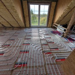 Instalacja ogrzewania podłogowego w nowym domu. Białe rurki ułożone na czerwonej macie z siatką, widok na okno i ocieplone poddasze. Etap przed wylewką.
