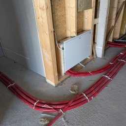 Czerwone rury ogrzewania podłogowego, przymocowane do rozdzielacza na ścianie w surowym wnętrzu. Widoczne ocieplenie ścian i płyty gipsowe.