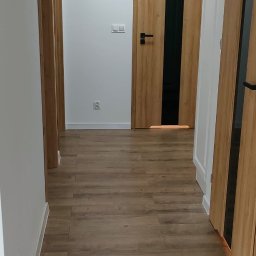 Edward Bobrowski - Korytarz po remoncie: panele podłogowe w odcieniach brązu, białe ściany z kontaktami, drewniane drzwi z czarnymi wstawkami i klamkami. Widok z perspektywy wejścia.