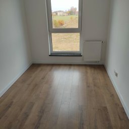 Edward Bobrowski - Jasne wnętrze z panelami podłogowymi w odcieniach brązu, białe ściany i okno z widokiem na zewnątrz. Grzejnik pod oknem. Minimalistyczny design.