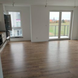 Edward Bobrowski - Jasne wnętrze z panelami podłogowymi w odcieniach brązu. Widok na balkon z przeszkloną balustradą i krajobraz za oknem. Częściowo widoczna zabudowa kuchenna.