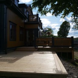 Edward Bobrowski - Jasny, drewniany taras przylegający do budynku z żółtą elewacją i ciemnymi detalami. Widoczne balustrady i elementy architektury. Błękitne niebo z chmurami w tle.