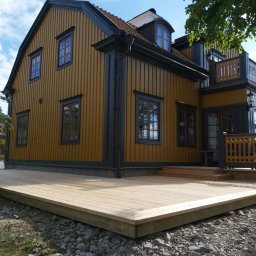 Edward Bobrowski - Żółty dom z drewnianą elewacją i nowym, jasnym tarasem z desek. Widok z boku, w tle błękitne niebo z chmurami. Kamieniste podłoże pod tarasem.