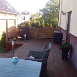 Edward Bobrowski - Drewniany taras z ażurową balustradą, meblami ogrodowymi i oświetleniem. Widok na dom i zieleń. Ujęcie z perspektywy, podkreślające przestrzeń i detale wykończenia.