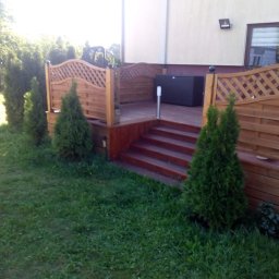 Edward Bobrowski - Drewniany taras z ażurową balustradą i schodami, otoczony zielonym trawnikiem i tujami. Na tarasie widoczny czarny pojemnik. Ujęcie z poziomu trawnika.