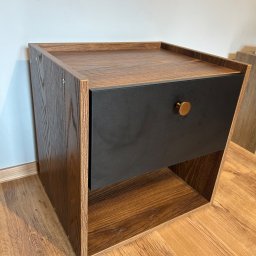 Szymon Szczygieł - Drewniana szafka nocna z czarną szufladą i złotym uchwytem, na tle ściany i podłogi z paneli. Widoczna faktura drewna i detale wykończenia.