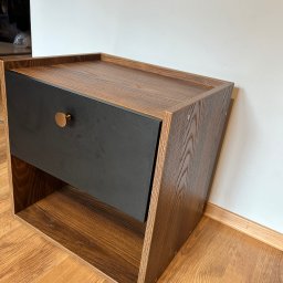 Szymon Szczygieł - Drewniana szafka nocna z czarną szufladą i złotym uchwytem, ustawiona na podłodze z paneli. Widoczne śruby mocujące elementy szafki.