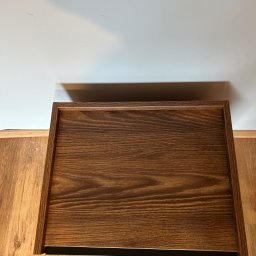 Szymon Szczygieł - Ciemnobrązowa, drewniana szuflada z metalowym uchwytem, widoczna z góry na tle podłogi i ściany. Wykończenie z wyraźnym rysunkiem słojów.