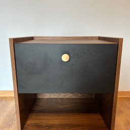 Szymon Szczygieł - Drewniana szafka nocna z czarną szufladą i okrągłym, złotym uchwytem, na tle białej ściany i drewnianej podłogi. Widok z przodu, minimalistyczny design.