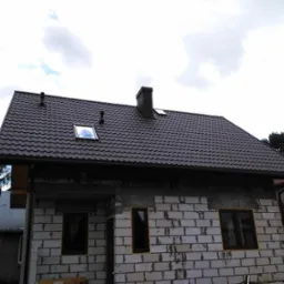 Nowy, ciemnobrązowy dach z dachówki trapezowej na surowym budynku z białego pustaka, widoczne okno dachowe i komin, w tle drzewa i sąsiednie domy.