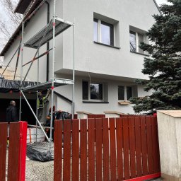 Mos-Bud - Prace elewacyjne na rusztowaniu przy dwukondygnacyjnym domu z szarą elewacją. Widoczny fragment czerwonego ogrodzenia i osoby w strojach roboczych.