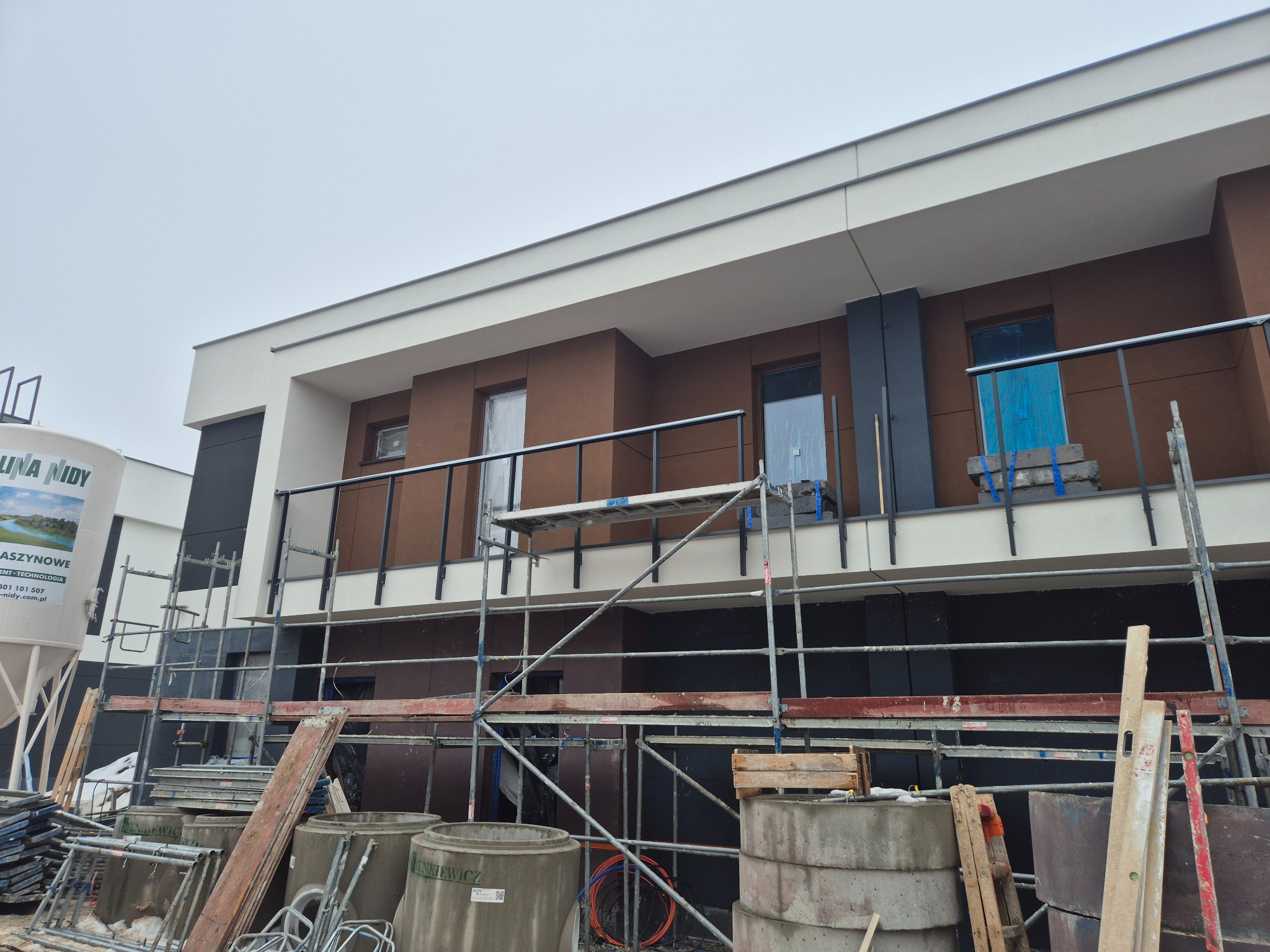 Nowoczesna elewacja budynku w trakcie prac wykończeniowych. Widoczne rusztowania, materiały budowlane i fragment zbiornika na zaprawę. Elewacja w kolorach brązu i bieli.