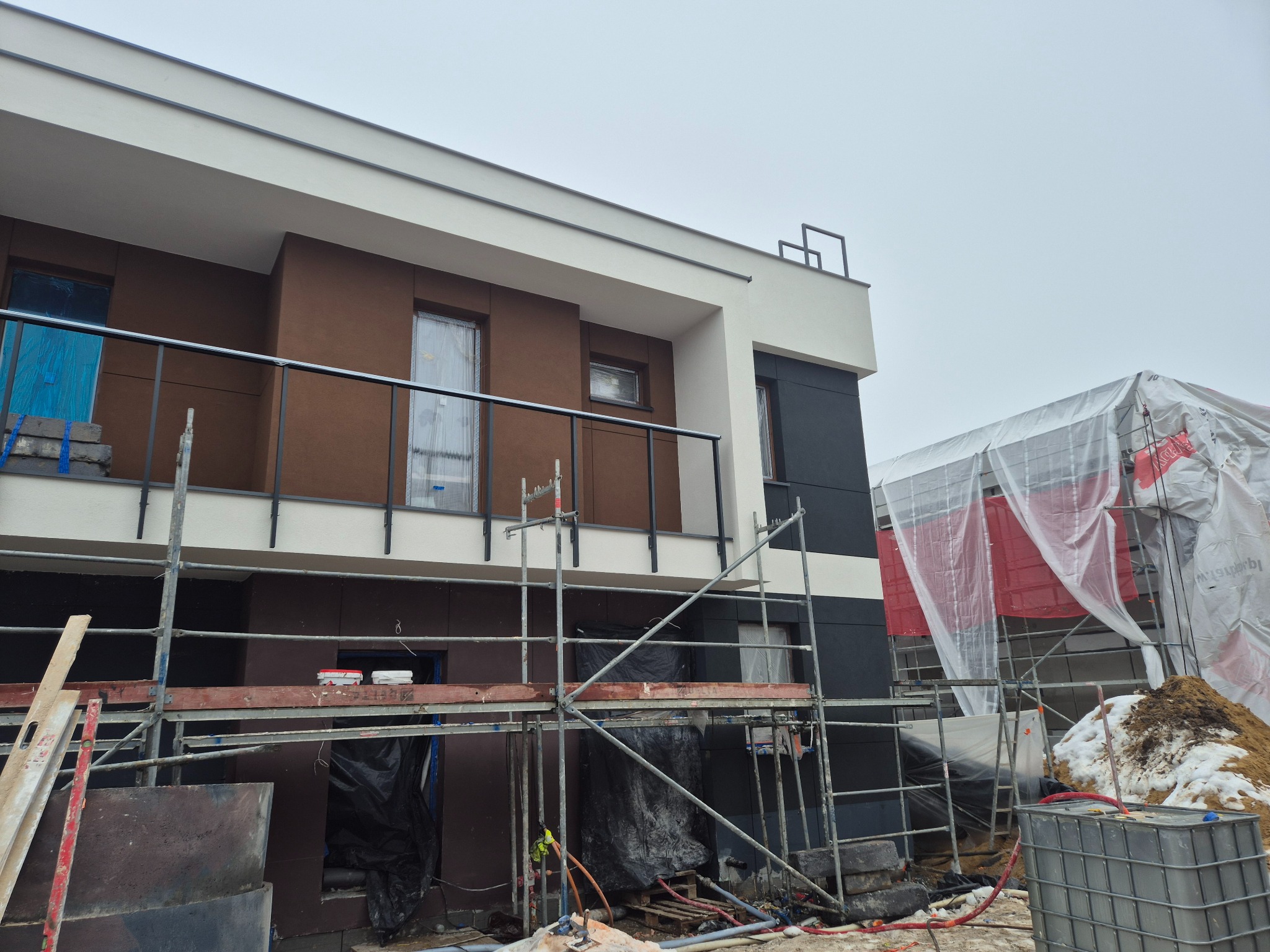 Dwukondygnacyjny dom w trakcie prac elewacyjnych: rusztowania, zabezpieczone okna folią, widoczne materiały budowlane. Część budynku osłonięta plandeką.