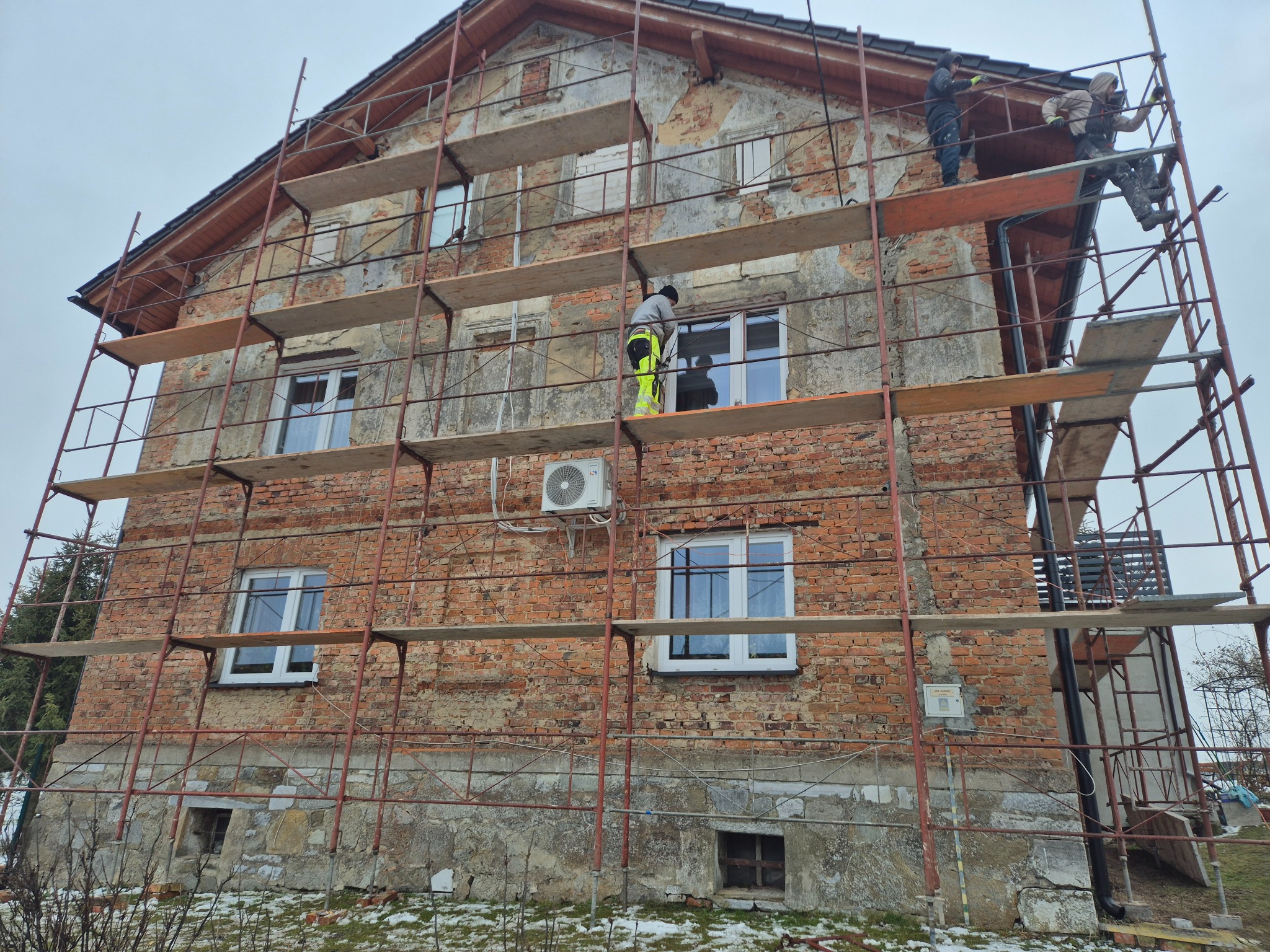 Dwupiętrowy dom z cegły otoczony rusztowaniem, na którym pracują robotnicy. Widoczne ślady starego tynku i przygotowania do prac elewacyjnych. Zima, pochmurne niebo.
