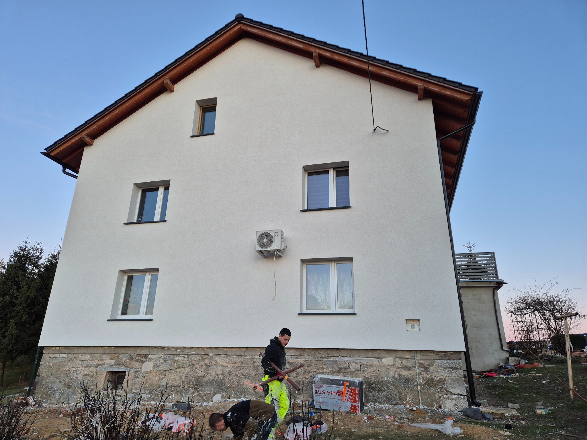 Dwóch pracowników przy ocieplaniu elewacji domu jednorodzinnego. Widoczna biała elewacja, styropian i narzędzia. Częściowo wykończona elewacja z klimatyzatorem.