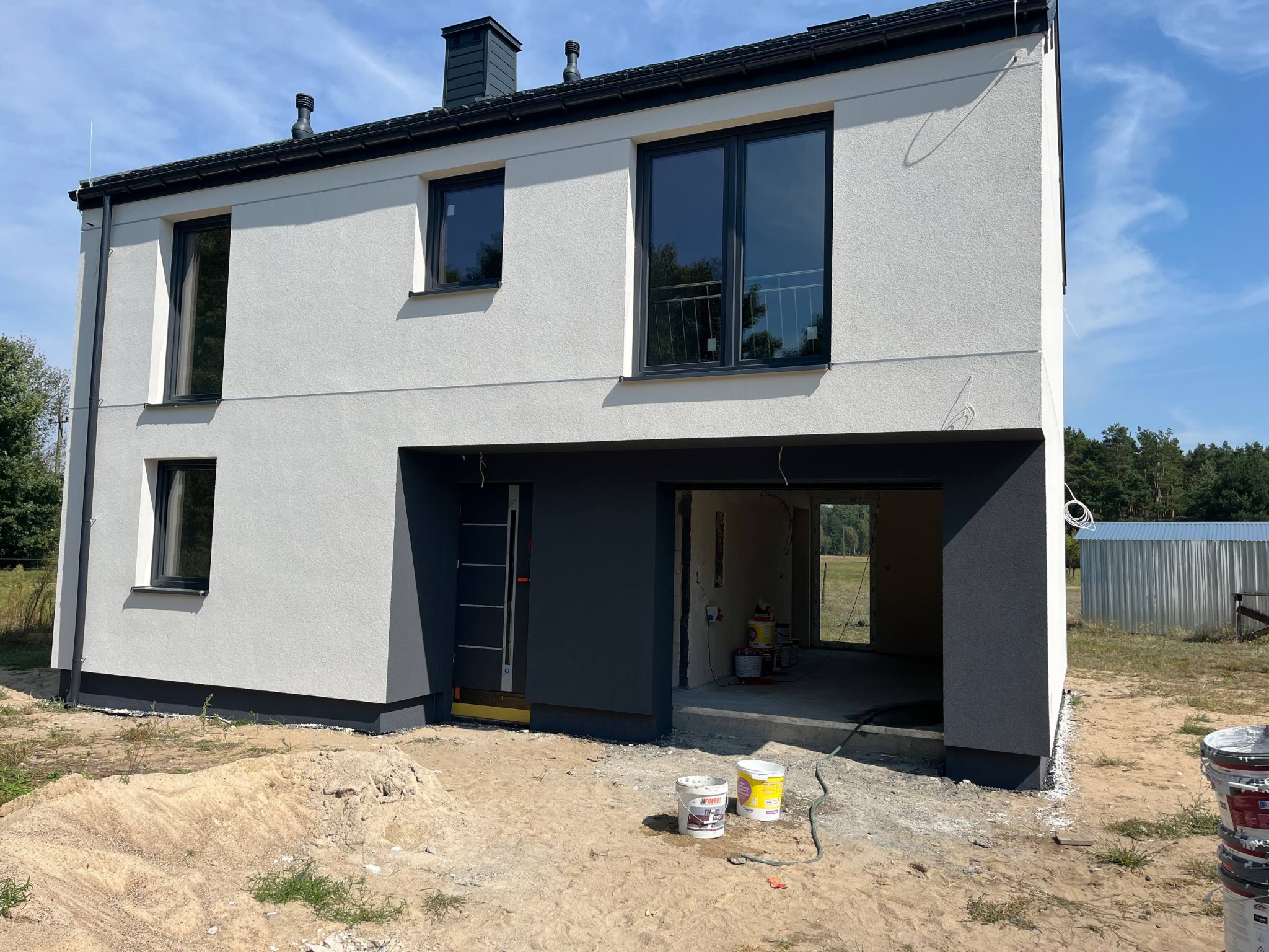 Dwukondygnacyjny dom z białą elewacją i antracytowymi detalami, widoczne puszki z farbą, sugerujące świeżo zakończone prace wykończeniowe.