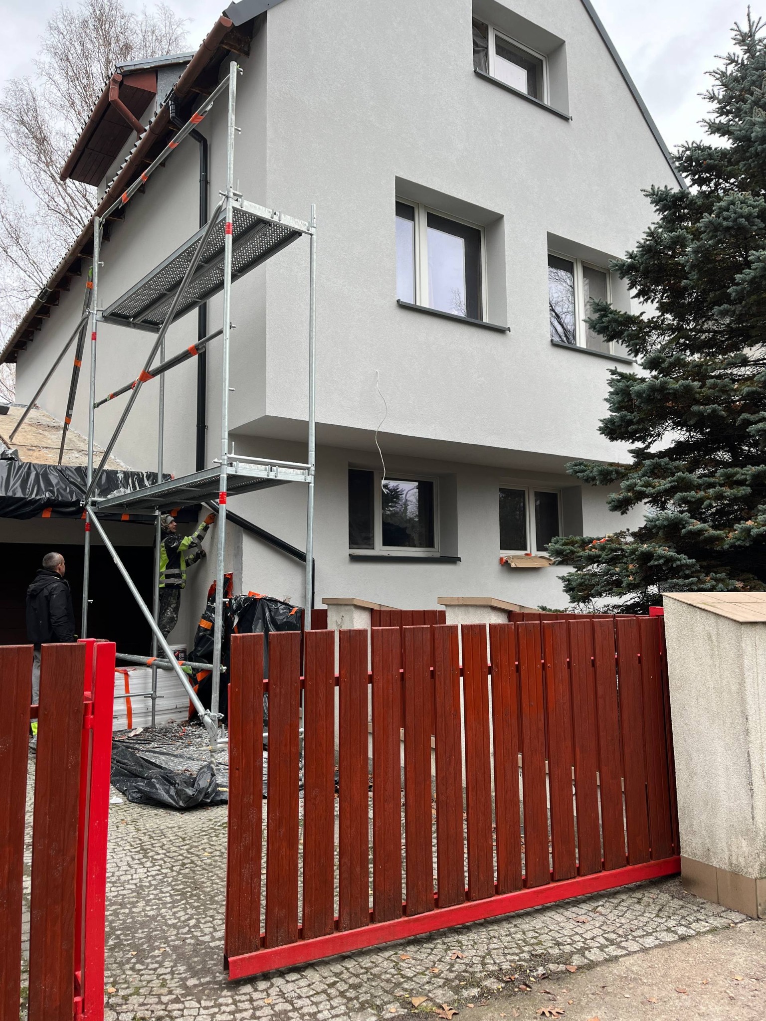 Prace elewacyjne na rusztowaniu przy dwukondygnacyjnym domu z szarą elewacją. Widoczny fragment czerwonego ogrodzenia i osoby w strojach roboczych.