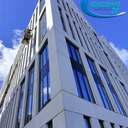 Aquacleaning24 - Wysoki budynek biurowy z pracownikiem na podnośniku koszowym podczas mycia okien. Widok z dołu, błękitne niebo w tle. Logo firmy w prawym górnym rogu.