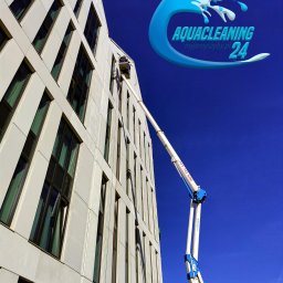 Aquacleaning24 - Wysokopodnośnik z pracownikiem przy elewacji budynku podczas mycia okien. Logo firmy Aquacleaning w prawym górnym rogu. Błękitne niebo w tle.
