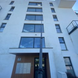 Aquacleaning24 - Mycie okien na wysokości w Gdańsku. Widok z dołu na białą elewację budynku z oknami, na tle błękitnego nieba, z użyciem specjalistycznego sprzętu do czyszczenia.