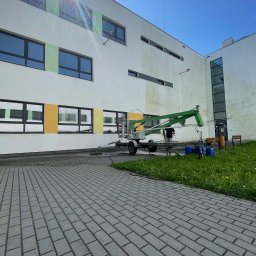 Aquacleaning24 - Elewacja budynku w trakcie czyszczenia z użyciem podnośnika i myjki ciśnieniowej. Widoczne ślady czyszczenia, kostka brukowa i trawnik.