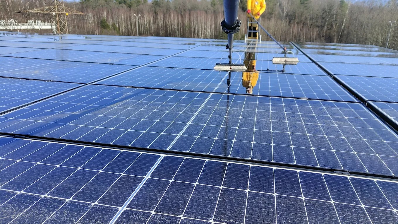 Czyszczenie paneli fotowoltaicznych z użyciem szczotki na wysięgniku. Widoczne odbicie pracownika w umytych panelach. W tle las i słup wysokiego napięcia.