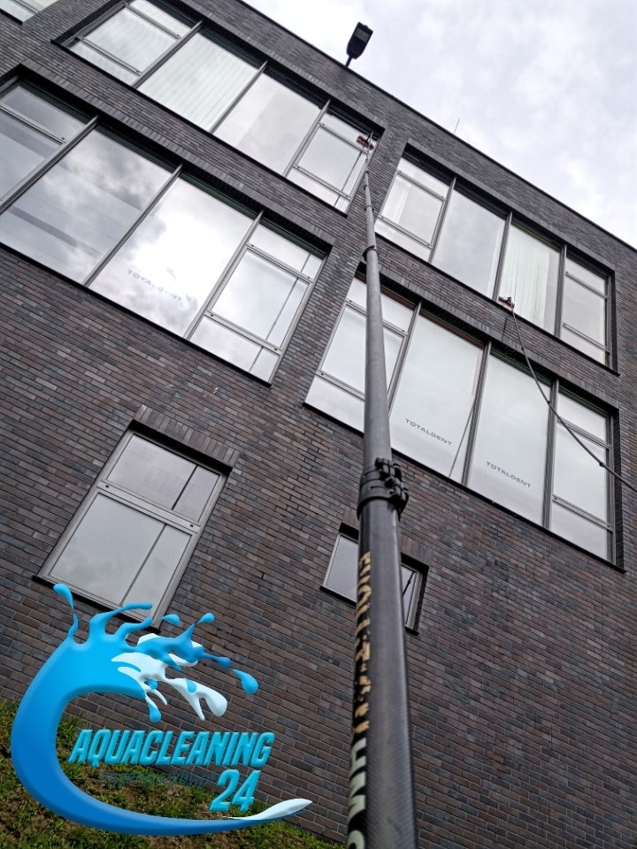 Mycie okien na wysokości z użyciem długiej lancy teleskopowej. Widoczna ceglana elewacja budynku z dużymi oknami. Na oknach widoczne logo firmy.