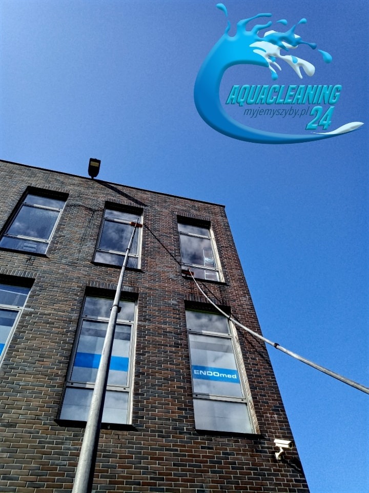 Czyszczenie okien na wysokości w budynku biurowym z cegły, za pomocą długiej lancy teleskopowej. Widoczne logo firmy Aquacleaning i niebieskie niebo.