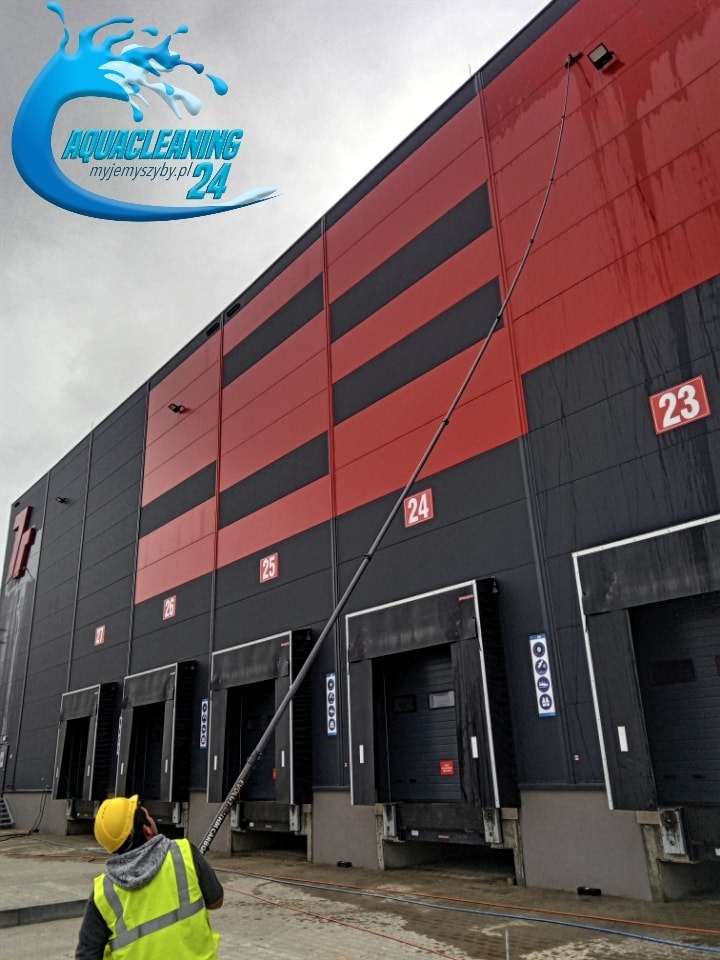 Pracownik w kasku myje elewację magazynu z użyciem długiej lancy teleskopowej. Widoczne numery bram, czarno-czerwona kolorystyka budynku i logo firmy.