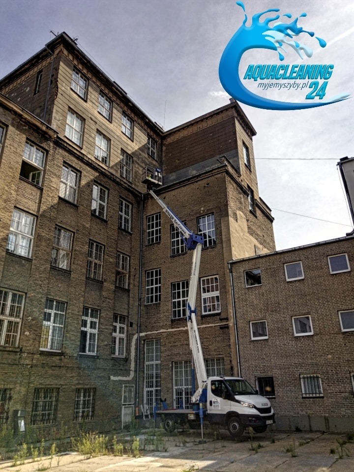 Wysięgnik koszowy przed budynkiem z cegły podczas prac na wysokości. Widoczny fragment elewacji i okna. Logo firmy w prawym górnym rogu.