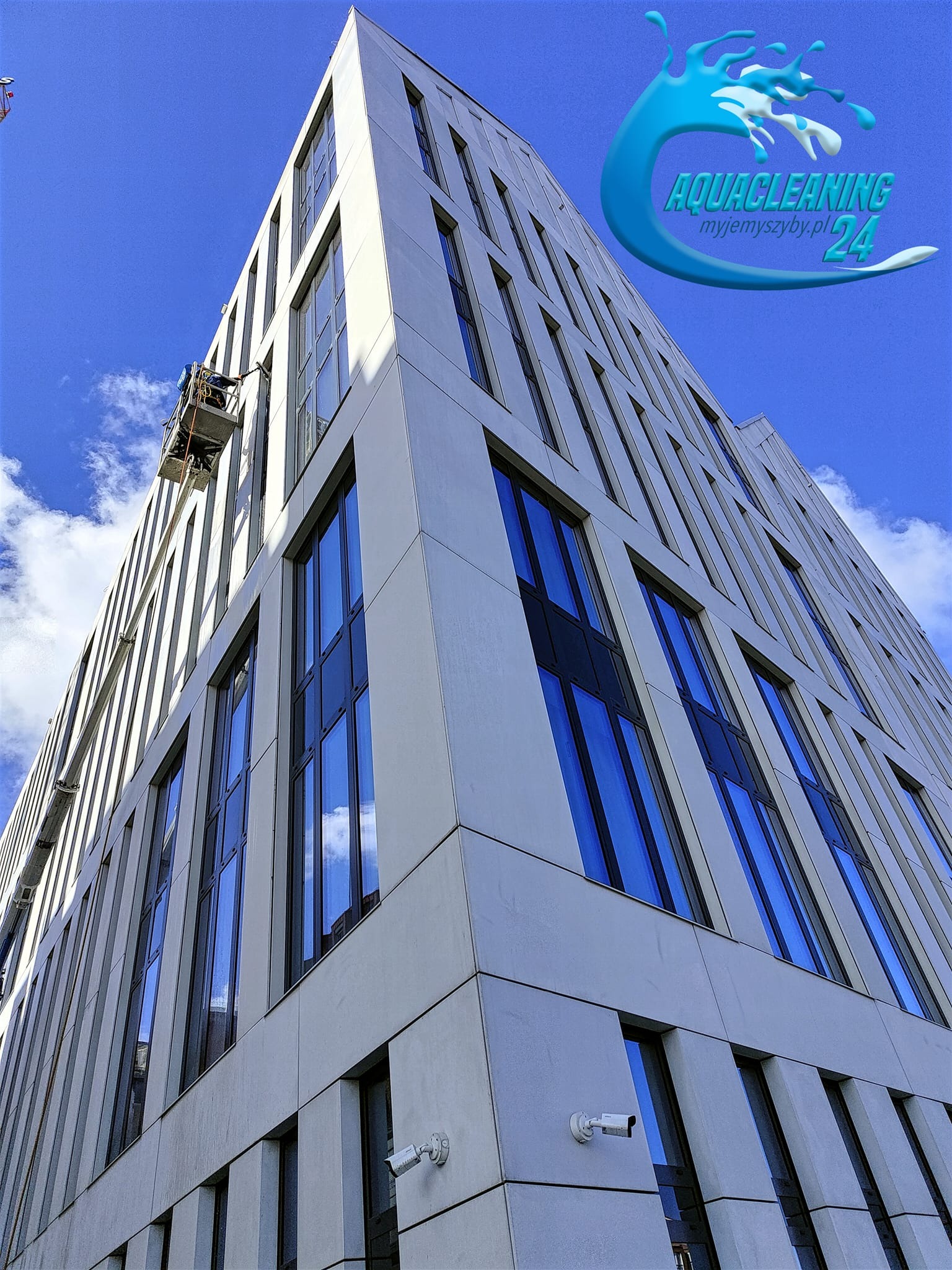Wysoki budynek biurowy z pracownikiem na podnośniku koszowym podczas mycia okien. Widok z dołu, błękitne niebo w tle. Logo firmy w prawym górnym rogu.