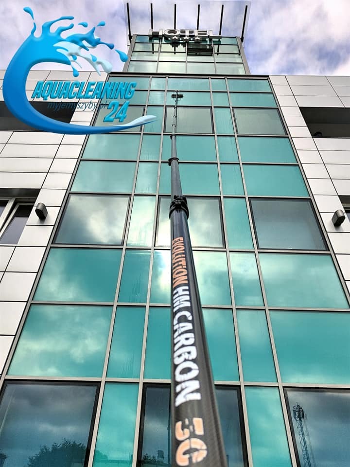 Mycie okien w hotelu za pomocą teleskopowej lancy z napisem 'EVOLUTION HM CARBON 5G'. W tle błękitne niebo odbijające się w szybach.