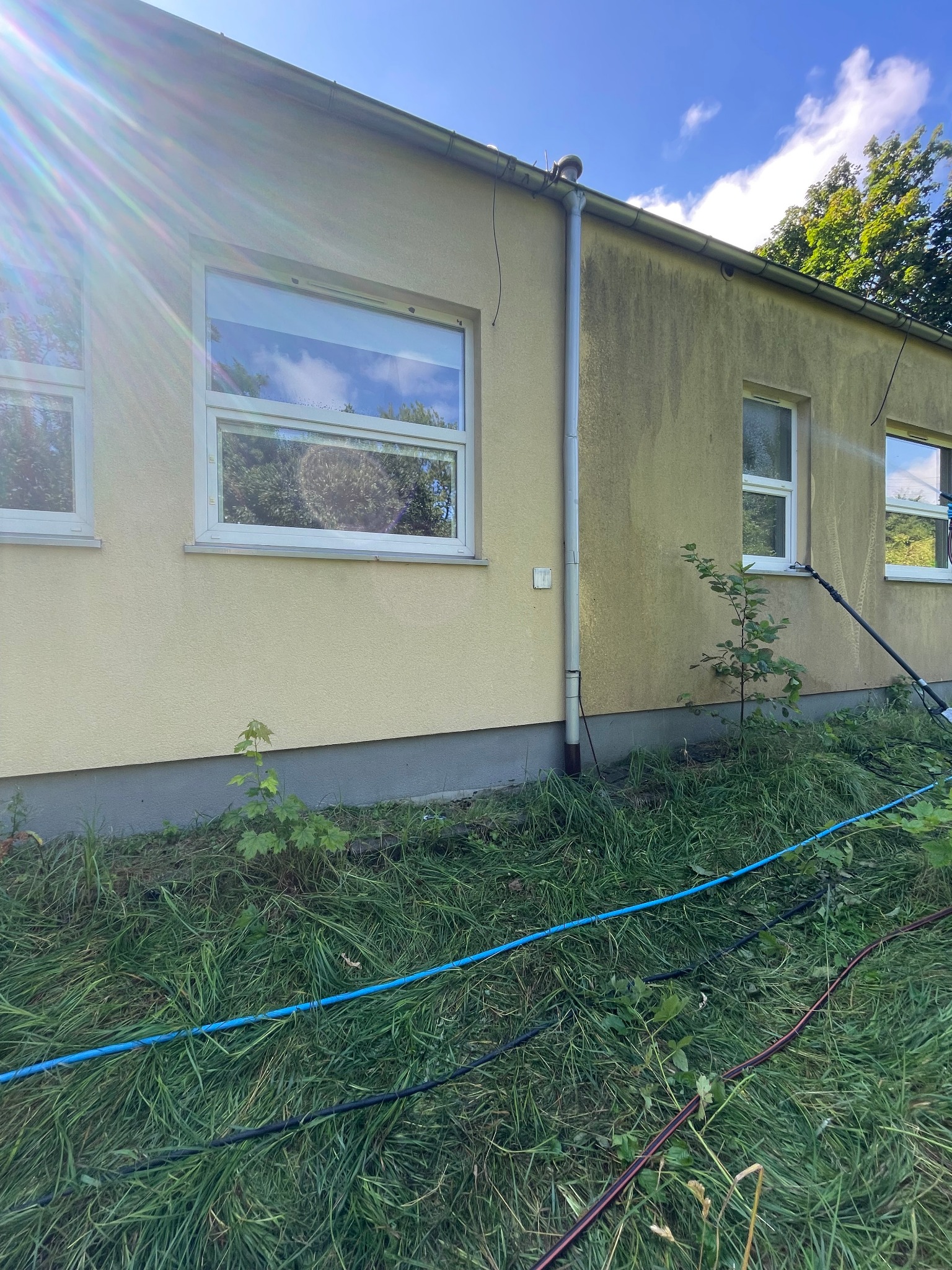 Żółta elewacja budynku z widocznymi zabrudzeniami, czyszczona lancą teleskopową. W tle zieleń drzew i błękit nieba. W trawie leżą węże doprowadzające wodę.