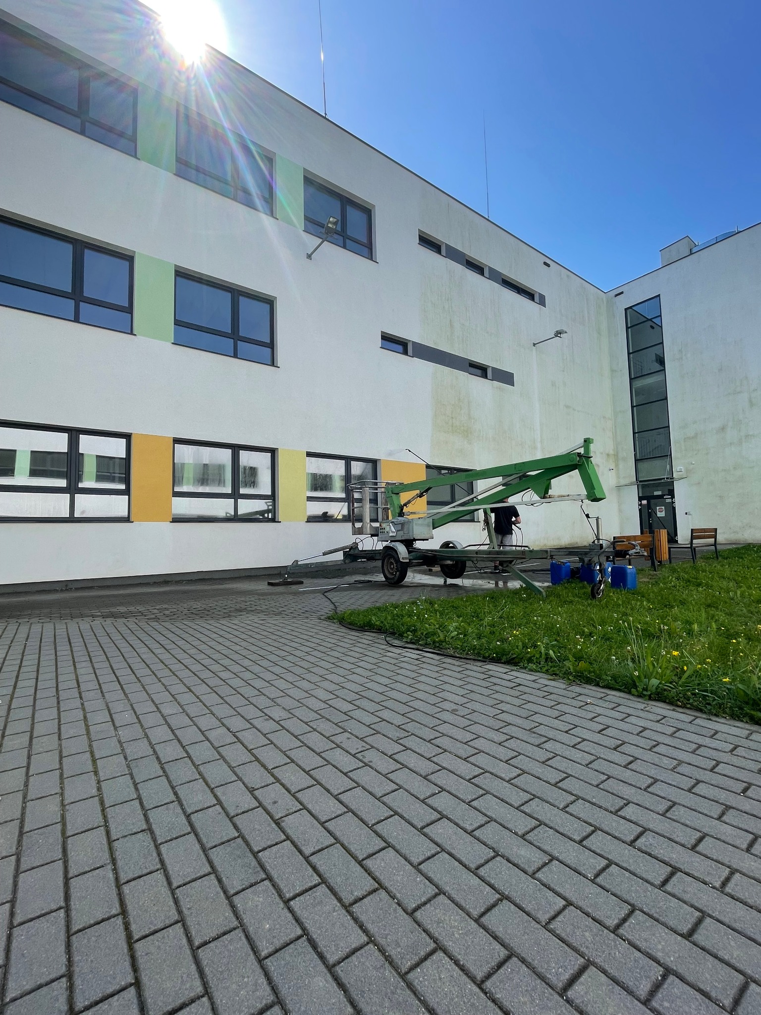 Elewacja budynku w trakcie czyszczenia z użyciem podnośnika i myjki ciśnieniowej. Widoczne ślady czyszczenia, kostka brukowa i trawnik.