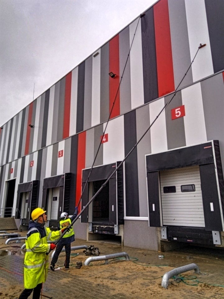 Dwóch pracowników w odblaskowych ubraniach myje elewację magazynu za pomocą długich lanc. Widoczne numery 4 i 5 na elewacji. Mokra nawierzchnia.