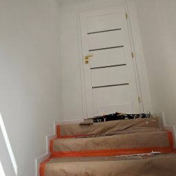 Wojciech Partyka - Klatka schodowa w trakcie zabezpieczania przed malowaniem: schody oklejone papierem i folią, białe ściany, białe drzwi z poziomymi paskami, lampa sufitowa.