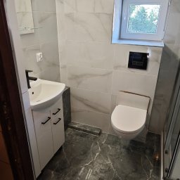 Wojciech Partyka - Nowoczesna łazienka po remoncie: białe marmurowe płytki ścienne, ciemna marmurowa podłoga, biała szafka z umywalką i czarną armaturą, okno i toaleta.