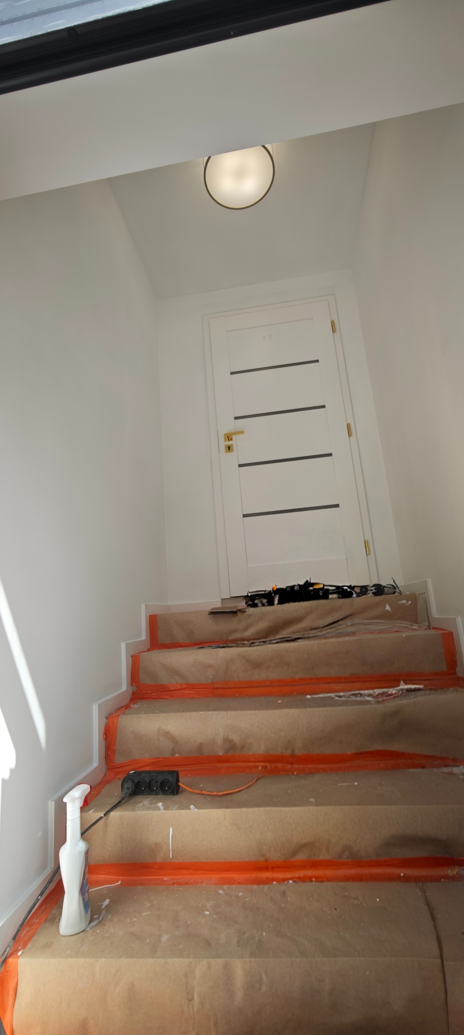 Klatka schodowa w trakcie zabezpieczania przed malowaniem: schody oklejone papierem i folią, białe ściany, białe drzwi z poziomymi paskami, lampa sufitowa.