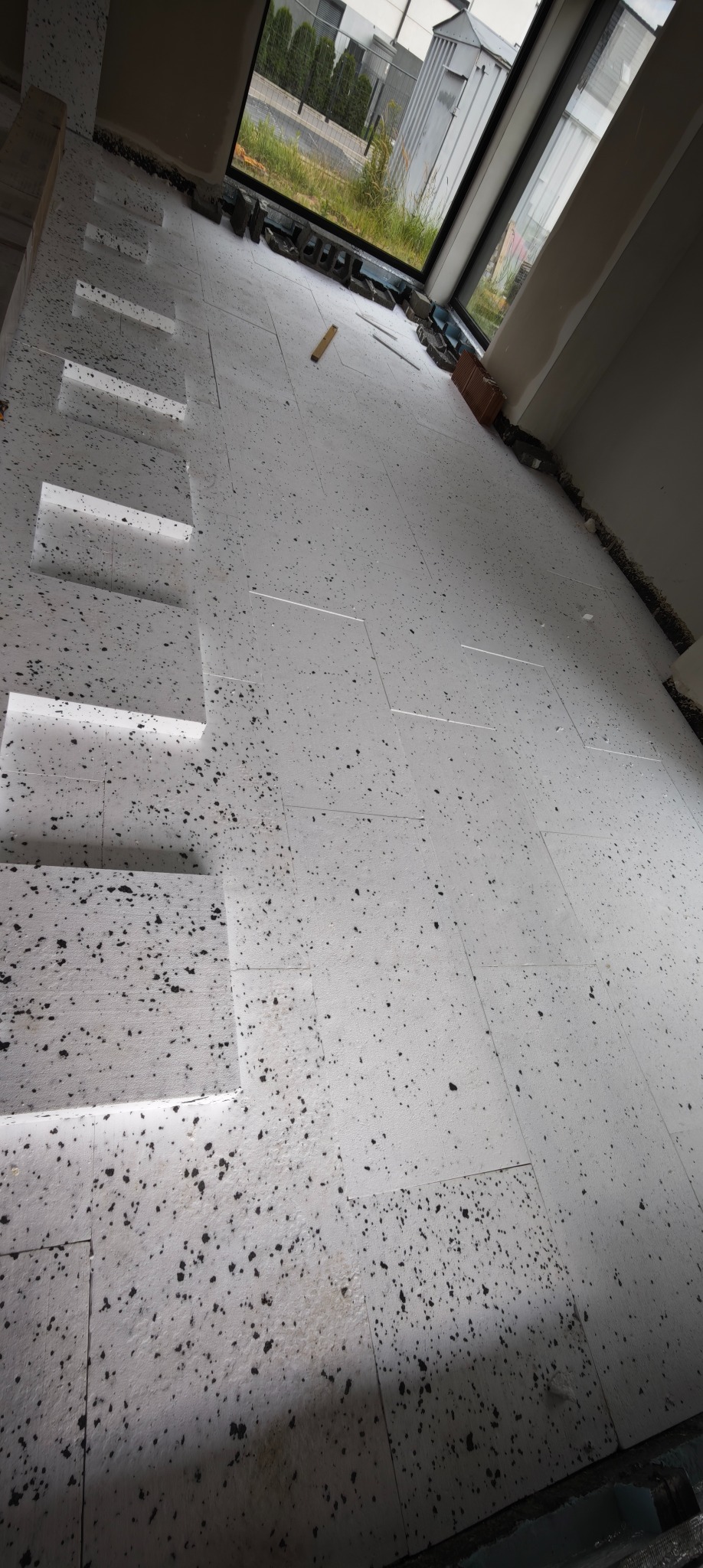 Układanie białego styropianu z czarnymi plamkami na podłodze w nowym budynku, przygotowanie pod posadzkę. Widoczne okno i fragment ściany.