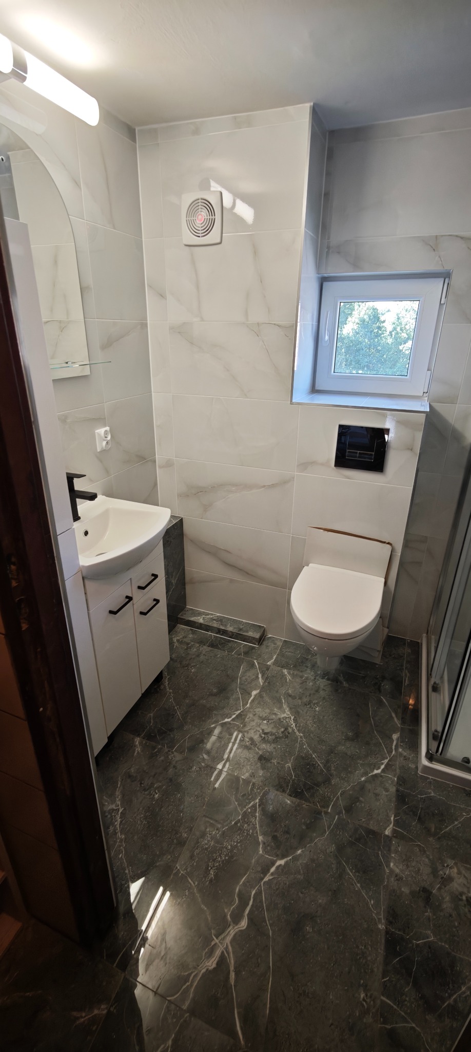 Nowoczesna łazienka po remoncie: białe marmurowe płytki ścienne, ciemna marmurowa podłoga, biała szafka z umywalką i czarną armaturą, okno i toaleta.