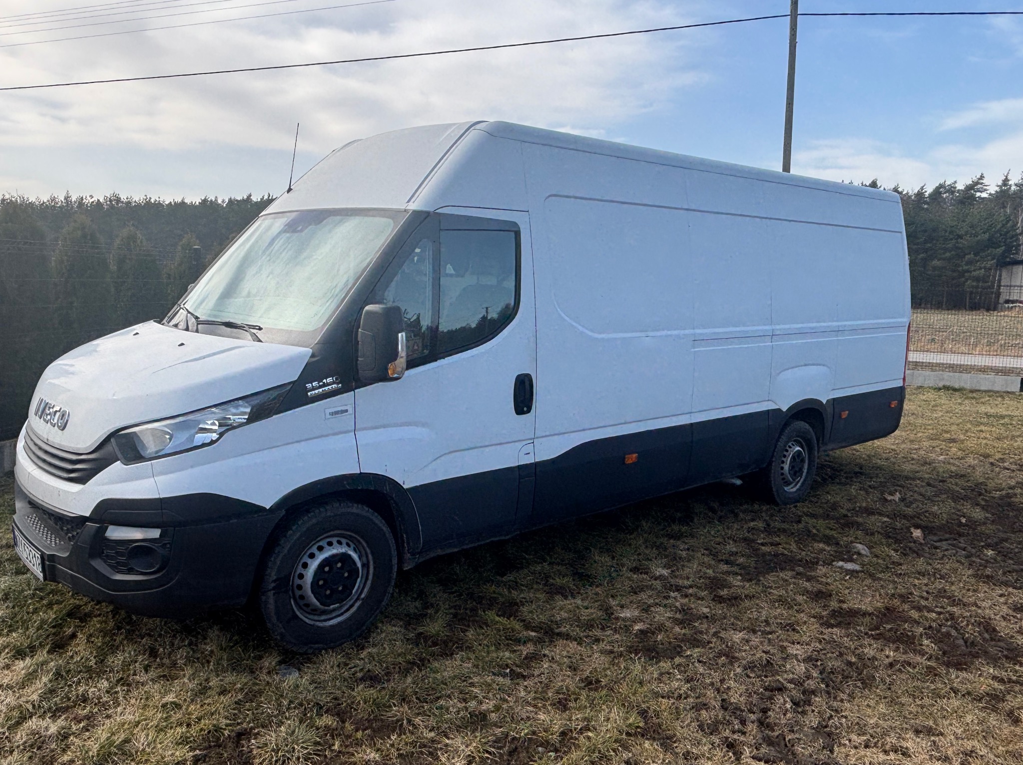 Biały furgon Iveco na trawiastym terenie, widok z boku, z drzewami w tle pod błękitnym niebem. Pojazd gotowy do transportu lub przeprowadzki.