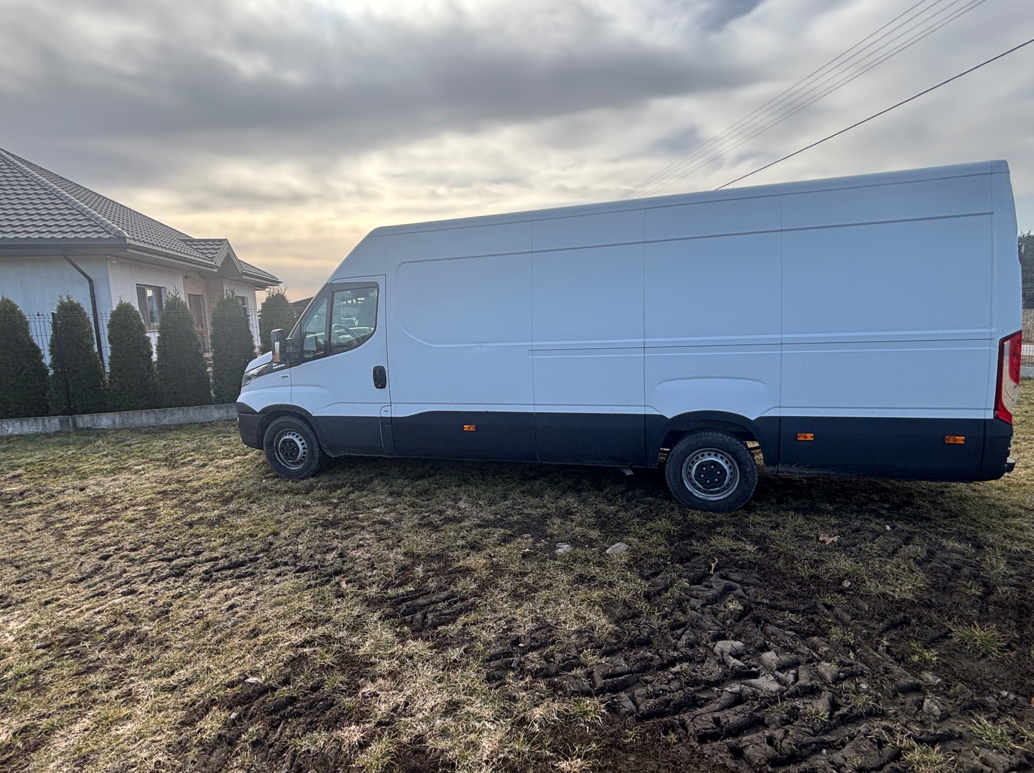 Biały bus dostawczy na błotnistym terenie przed domem z brązowym dachem w pochmurny dzień. Widoczne ślady opon na trawie i fragment linii energetycznej.