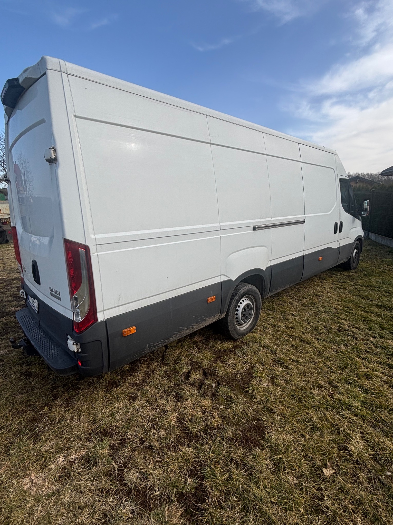 Biały, długi bus dostawczy na trawiastym terenie, widziany z perspektywy tylnej, lekko z boku. Widoczne detale karoserii i opon. Błękitne niebo w tle.