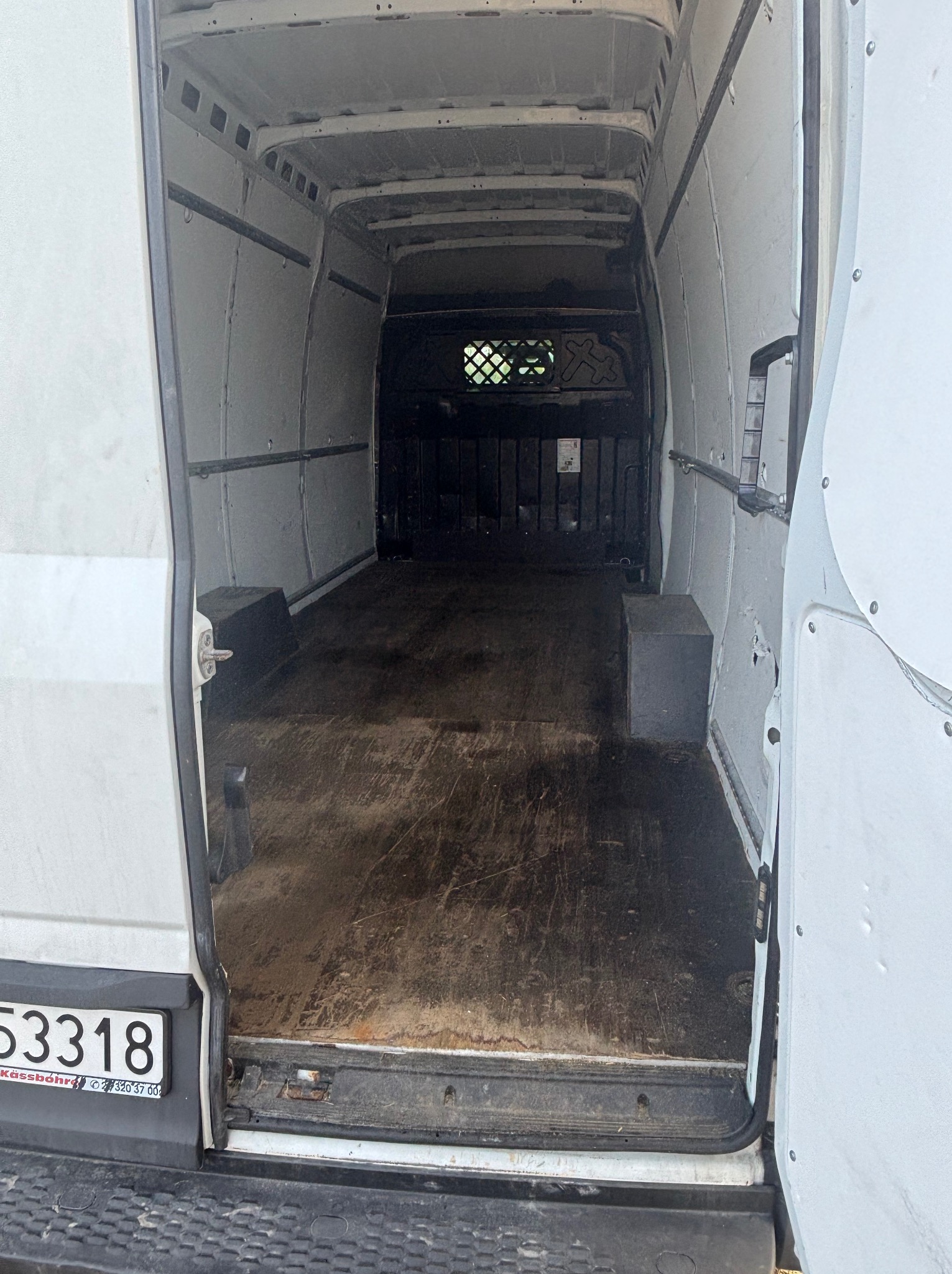 Wnętrze białego busa z otwartymi drzwiami, widoczna pusta przestrzeń ładunkowa z drewnianą podłogą. Tablica rejestracyjna widoczna w lewym dolnym rogu.