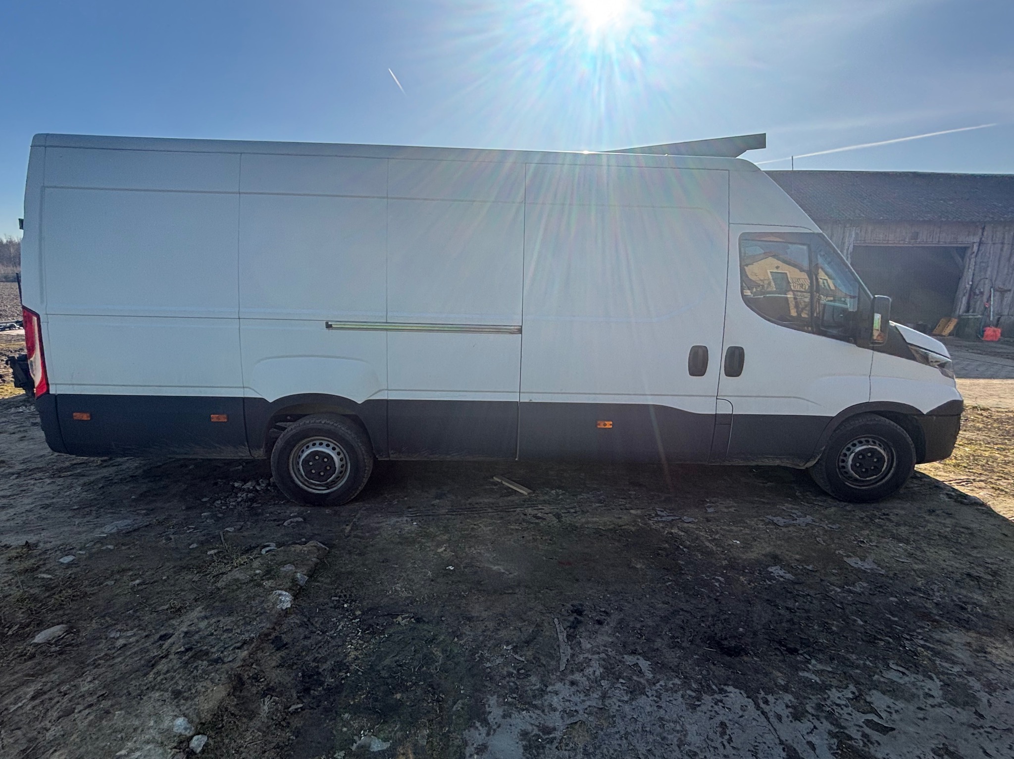 Biały bus dostawczy na błotnistym podłożu, widok z boku, jasne słońce w tle. Charakterystyczny cień pod pojazdem. Budynek gospodarczy w tle.