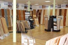 Wnętrze sklepu z ekspozycją różnorodnych paneli podłogowych i desek drewnianych, ułożonych pionowo na stojakach i w rzędach wzdłuż ścian, oświetlone światłem dziennym i sztucznym.