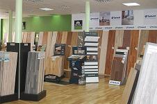 Ekspozycja w sklepie z szerokim wyborem wzorów i odcieni paneli podłogowych i desek, z widocznymi stojakami prezentacyjnymi i plakatami reklamowymi producentów.