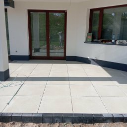 Krzysztof Roguski - Jasne płyty tarasowe ułożone na zewnątrz budynku z ciemną podmurówką i oknami. Widoczne fugi i obrzeża z kostki. W tle odbicie osoby w oknie.