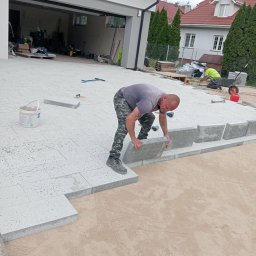 Krzysztof Roguski - Pracownik w trakcie układania jasnoszarej kostki brukowej na podsypce piaskowej. W tle widoczny budynek mieszkalny i garaż z otwartą bramą.